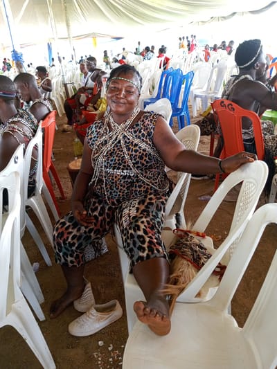 The image shows a woman seated in a white plastic chair under a white tent in Takau, Nigeria. She is wearing leopard-print pants and a top, and multiple strands of white beads around her neck. She has a beaded headband. One foot is bare, resting on another chair, with her shoes on the ground nearby. Other people, some in similar attire with beaded necklaces and headbands, are seated in the background in white, blue, and orange plastic chairs. A water bottle is visible next to a wooden drum. The ground appears to be dirt.