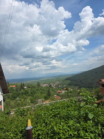 This panoramic image presents a picturesque rural landscape, likely captured from an elevated position overlooking a valley. The scene depicts a bright, partly cloudy day over Slokoshtitsa, Bulgaria.
In the foreground, at the bottom of the frame, dense green foliage, possibly from bushes or garden plants, creates a natural border. Elements of a building, including a wooden wall, a white structure, and a brown-tiled roof, are visible at the bottom edge, suggesting the viewpoint is from a property. A yellow-tipped pole stands prominently amidst the greenery on the right.
The midground is dominated by a charming village or settlement nestled in the valley. Numerous houses with characteristic red-tiled roofs are scattered amidst a rich tapestry of green trees and cultivated fields, indicating a vibrant rural community.
Stretching across the background is a series of rolling hills and mountains. The nearest range is densely forested with dark green trees, forming a distinct horizon. Beyond this, further hazy mountain ranges recede into the distance, contributing to the expansive feel of the landscape.
The sky, occupying the upper portion of the image, is a bright blue canvas adorned with large, fluffy white cumulus clouds. Some clouds appear thicker with grey undersides, adding depth to the atmospheric conditions. Several thin utility wires or power lines are visible against the sky in the upper left section. The overall impression is one of serene natural beauty and rural tranquility under clear daytime conditions. There are no people or specific activities depicted, and no visible text.