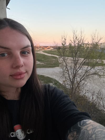 The image is a selfie of a young woman taken outdoors in Chornomorskoe, Ukraine, during what appears to be either sunset or sunrise. The woman, who is the primary subject, is positioned in the foreground, looking directly at the camera with a neutral to slightly serene expression. She has long dark hair, blue eyes, fair skin with some freckles, and wears a septum piercing. Her lips are full and slightly parted.

She is dressed in a dark, likely black, long-sleeved shirt. On her shirt, near her right shoulder (visible on the right side of the image), there is a white and red-capped character resembling an "Among Us" crewmate, with partial text "adi" visible beside it, suggesting an Adidas brand. Her right arm, which is partially visible, is covered in extensive, intricate tattoos. A light-colored concrete or stone surface is visible in the bottom-left corner, possibly a ledge she is leaning against.

The background reveals a transitional landscape with dirt paths winding through areas of grass and bare earth. Several trees, some with sparse green leaves budding and others still mostly bare, are scattered across the mid-ground. In the far distance, buildings indicating a town or settlement are visible. The sky transitions from a soft light blue on the left to delicate pink and orange tones on the right, characteristic of dawn or dusk. The overall setting suggests a somewhat rural or suburban periphery of Chornomorskoe.