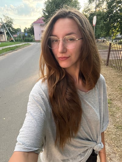 The media file is a selfie taken outdoors in Zhodzina, Belarus, with the camera held in a vertical orientation. The subject is a young woman with long, brown hair and glasses. She is wearing a light gray, short-sleeved t-shirt that is tied in a knot at her side. The background shows a paved road, a grassy area with dry vegetation, a metal fence, and trees. In the distance, a few houses and street signs are visible. The lighting suggests it is either early morning or late afternoon, with the sun casting a warm glow. The sky is partly cloudy. The woman's expression is relaxed and she is looking towards the camera. There is no visible text in the media file.