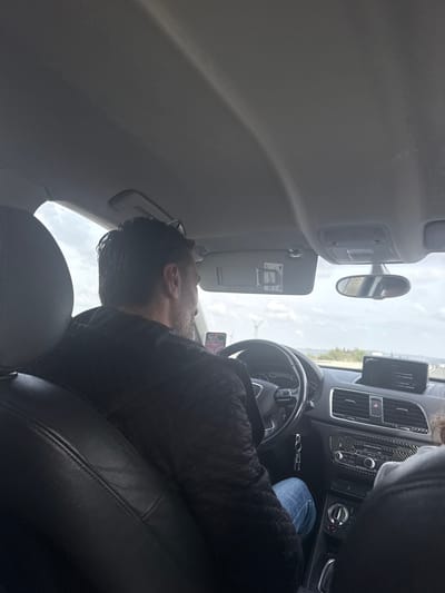 The scene depicts the interior of an Audi Q3 vehicle in motion, viewed from a rear passenger seat, within the city of Luc-sur-Orbieu, France. A male driver, with dark hair and a beard, wearing a dark, textured jacket and blue jeans, is seated at the steering wheel, looking forward. His left hand is visible near the steering wheel. A mobile phone, displaying what appears to be a navigation interface, is mounted to the left of the steering wheel. A set of keys hangs below the steering column. The dashboard features an infotainment screen displaying text, air vents with carbon fiber trim, and climate controls. Partially visible in the front passenger seat is a second individual, indicated by a headrest and a grey-clad arm or shoulder. The vehicle's headliner is light grey, and the seats are black leather. Outside the vehicle, the sky is overcast, and multiple wind turbines are visible in the distance above a line of vegetation, suggesting an open landscape.