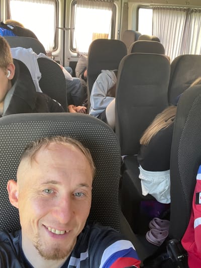 A selfie taken from inside a passenger van shows a man with short, light brown hair and a goatee smiling. He is wearing a black t-shirt with a white and blue stripe design on the sleeve. The interior of the van is visible behind him, with rows of dark-colored seats. Some passengers are seated in the background, some appear to be sleeping or resting. A white t-shirt is draped over a seat. Curtains cover the windows. There are also a pair of pink Crocs visible in the footwell of the van. The scene suggests a journey or transportation within a populated area.