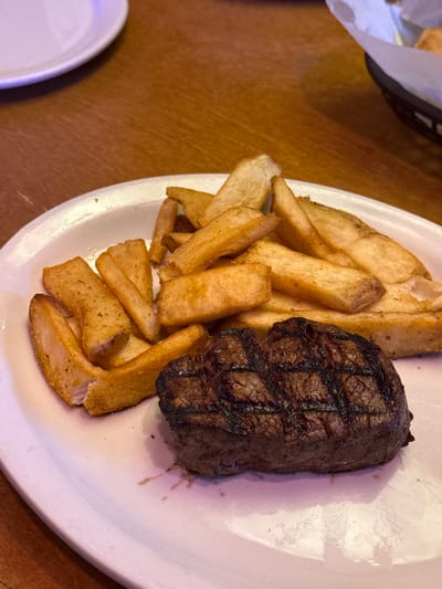 The image shows a plate of food. On the plate are french fries and a piece of grilled meat, likely steak. The plate sits on a wooden table. Part of another plate and a basket lined with paper are also visible in the image. The location is unknown.