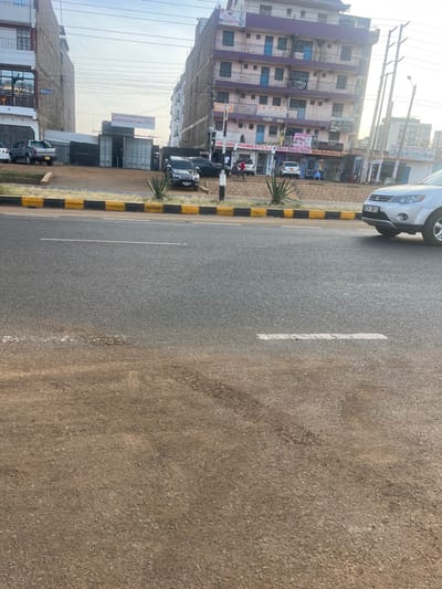The image displays an urban street scene in Gitothua ward, Kenya, characterized by a multi-lane asphalt road, commercial buildings, and vehicles under a bright, hazy sky.
In the foreground, an unpaved dirt shoulder transitions to a two-lane asphalt road, marked by a white dashed line. A yellow and black painted concrete median separates the visible lanes from oncoming traffic or a central reservation. Beyond the median, an elevated dirt strip with sparse agave-like plants runs parallel to the road.
To the left, a multi-story building with white or light-colored facades is visible. Adjacent to it, an open lot features several grey shipping container structures, one bearing a sign for "PEATON HOMES HARDWARE." A grey pickup truck is parked within this lot.
To the right, a prominent multi-story building, approximately six to seven stories tall, dominates the background. It features a light pink/purple exterior with blue window frames or railings on its upper floors. The ground floor houses multiple businesses, including "JUMIA O" (a Jumia online pick-up point), "HAFWAK PHARMACEUTICALS," and "Shera Beauty Salon." Several vehicles, including a black SUV and a dark sedan, are parked directly in front of these establishments. Approximately five individuals are observed walking or standing on the sidewalk near these businesses.
Overhead, a network of utility wires and poles is visible against the light sky. On the far right, the front portion of a silver SUV (license plate partially visible as KCY 581U) is captured on the asphalt road, suggesting movement or recent parking.
