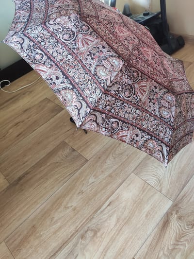 The image shows a close-up, high-angle view of a partially opened umbrella with a paisley-like pattern in shades of maroon, pink, white, and brown. The umbrella is lying on a light brown wooden floor. To the left of the umbrella, partially visible, is the top of a black vacuum cleaner with a hose. The scene is indoors, with the wooden floor suggesting a home or a similar interior space. The lighting appears to be natural or from an overhead source, casting soft shadows. No people are visible in the image. The overall impression is one of domestic clutter, with the umbrella and vacuum cleaner suggesting recent use or storage. The pattern on the umbrella is intricate and detailed, featuring floral and curved motifs. The wooden floor has a visible grain and appears to be laminate or wood flooring. There is a white cord extending from the umbrella towards the bottom right of the frame, which might be part of its mechanism or a separate item.