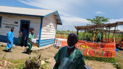 This image depicts an outdoor medical facility in Tchere, Democratic Republic of the Congo, under a bright, sunny sky. In the immediate foreground, a bald man wearing green medical scrubs, a white apron, and a surgical mask pulled down around his chin looks towards the camera.

Behind him, to the left, stands a simple, single-story building constructed from white wooden planks with blue trim and a corrugated metal roof. Signs on the building identify it as "FH RDC," "SOFEPAD," and "HOPITAL GENERALE DE REFERENCE RWAMPARA ESPACE SUR," indicating it is the Rwampara General Reference Hospital's "Safe Space." Around the building's entrance, several people are engaged in activity: a man in a blue full-body protective suit stands to the left, while another in green scrubs carries a white box from the doorway. Two other individuals are partially visible inside the door.

To the right of the building, an open-air structure with a wooden frame and a light-colored tarp roof is visible. This area is enclosed by bright orange plastic safety fencing. Inside, two individuals, also in medical scrubs, masks, and gloves, appear to be working at tables. Several white plastic chairs are scattered near the fencing. The ground is a mix of dirt and sparse vegetation, with a tree stump prominent in the foreground. The scene suggests a busy, functional medical or public health operation, possibly a temporary clinic or triage area. A watermark "Joseph Kisuki" is visible in the lower right corner.