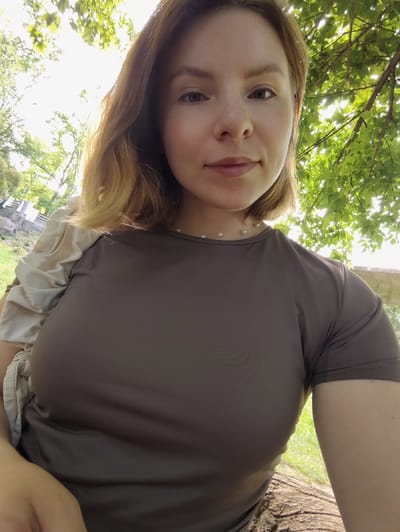 A selfie shows a Caucasian woman in her late 20s or early 30s, with shoulder-length blonde hair and blue eyes, wearing a brown t-shirt and a pearl necklace. She is outdoors, with green foliage and sunlight filtering through the trees behind her. The background suggests a park or natural setting, with a stone structure and grass visible to the left. The woman is seated, with a log in the foreground. The lighting is bright, with the sun in the upper left portion of the frame. The image does not contain any specific identifiable landmarks of Korosten, Ukraine.