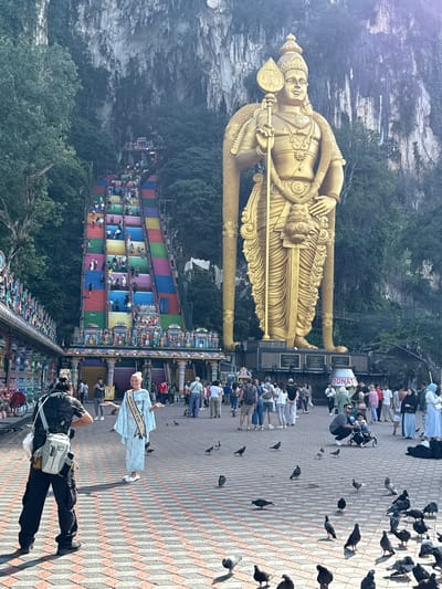 This outdoor photograph captures a bustling scene at the Batu Caves in Malaysia, featuring the prominent golden statue of Lord Murugan, a vibrant multi-colored staircase, and numerous people. The image is set under bright daylight, casting clear shadows on the tiled ground.

In the foreground, a large, diamond-patterned paved area is filled with a crowd of visitors and a multitude of pigeons, some perched, others walking. A striking figure in a light blue dress with a sash reading "MISS PETITE GLOBAL UK 2024" stands in the mid-ground. Nearby, a person dressed in dark clothing with a white shoulder bag is seen holding a camera, possibly photographing the woman in the sash or the broader scene.

Dominating the left side of the image is the colossal golden statue of Lord Murugan, depicted holding a spear (vel). This towering religious figure stands against a backdrop of a sheer limestone cliff face, partially covered by rich green foliage. Adjacent to the statue's base, a small structure with the visible word "DONATE" indicates a collection point.

To the right of the statue and extending into the background, a long, steep staircase ascends the hillside towards the cave entrance. Each step of this famous staircase is painted a different vibrant color, creating a rainbow effect that includes red, orange, yellow, green, blue, and purple hues. Many people are seen climbing and descending these stairs, highlighting the site's popularity as both a tourist attraction and a pilgrimage destination. The natural ruggedness of the limestone cliffs and surrounding trees provide a dramatic setting for the man-made structures.