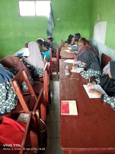 A classroom setting shows children at their desks. Some wear headscarves. They appear to be writing on paper. The desks are wooden, and a book rests on one of them. A window provides light on the left side. The walls are green with some white areas. Some students have backpacks. The scene suggests a focused activity in an educational environment.