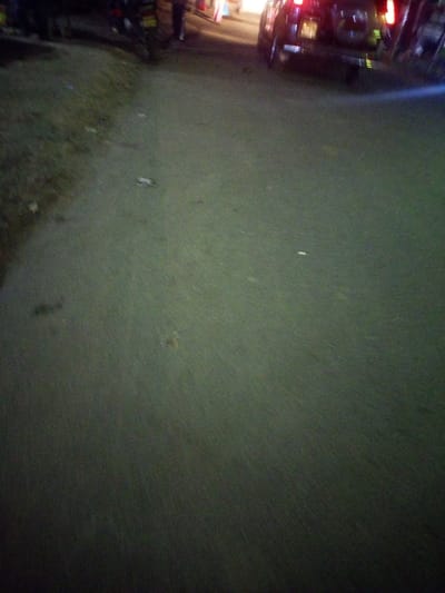 An asphalt road stretches into the distance under dim lighting. To the left, there's a grassy verge. A parked car with its taillights illuminated and a motorcycle are partially visible on the right, along with indistinct figures of people. The road itself is unevenly lit, with subtle variations in texture. A few small, light-colored objects are scattered on the road surface. The overall atmosphere is one of a dimly lit street scene at night.