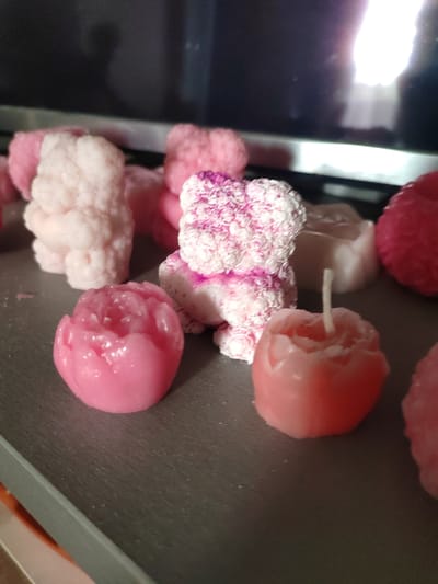 This media file displays an assortment of small, decorative candles in various pink and white hues. The subjects are primarily the candles themselves, which are shaped like flowers and a teddy bear. There are at least three flower-shaped candles, one distinctly resembling a peony, and a central candle shaped like a teddy bear, with pink speckled detailing on its white body. Other candles are partially visible, some appearing as amorphous shapes. The objects are placed on a dark, textured surface, possibly a tray or table. The background is dark and out of focus, with reflections suggesting it might be a screen or a window. There are no people visible, nor any indication of a specific time of day or weather conditions. The overall impression is that of a collection of artisanal or novelty candles. No text is visible in the media file.