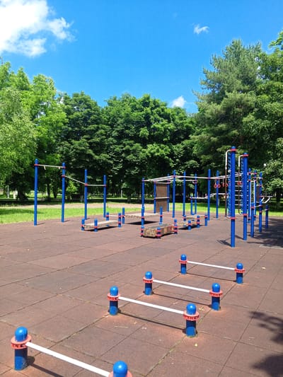 This is an outdoor photograph of a calisthenics park in Yessentukskaya, Russia. The park features a variety of exercise equipment, including parallel bars, pull-up bars, and an agility ladder, all constructed with blue metal poles and white bars. The ground is covered with red rubber safety mats, laid out in a grid pattern. Lush green trees form a dense backdrop to the park, and the sky above is a clear blue with a few scattered white clouds. The scene is devoid of people, suggesting it is either early in the morning, late in the evening, or during a period when the park is not in use. The overall impression is one of a well-maintained public space designed for outdoor physical activity.
