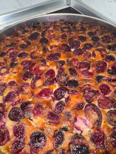 The image shows a close-up view of a cherry clafoutis in a metal baking pan. The clafoutis is richly studded with dark red and black cherries, baked into a custard-like batter that is golden-brown in places. The dessert appears to be freshly baked and is sitting on a metal surface, likely a kitchen counter or serving area. The location is Elhovo, Bulgaria.