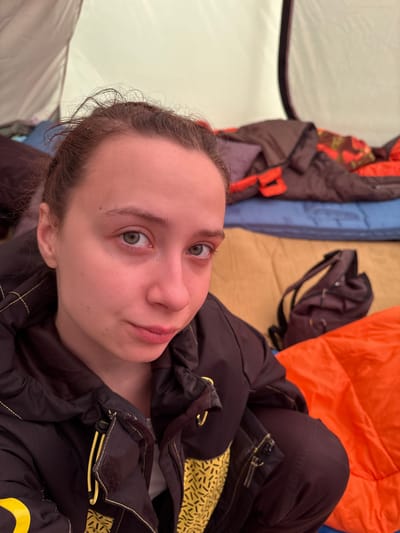 A fair-skinned person with light brown hair pulled back is lying down, looking up at the camera. The person is wearing a dark jacket with yellow patterned lining. The background consists of camping gear including sleeping bags in colors orange, brown, and blue. Part of a tent wall is visible on the left. The location is unspecified; there is no information to identify Lesnoye, Russia.
