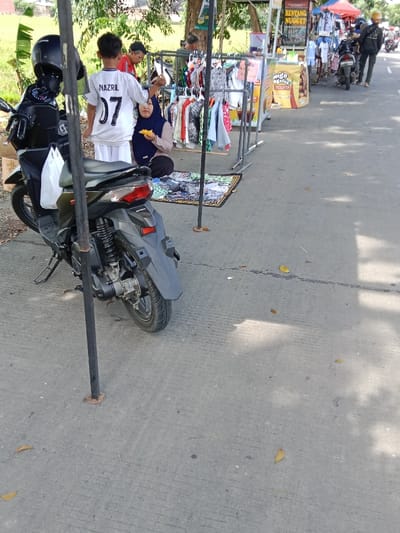 A black scooter is parked on the sidewalk in Kendal, Indonesia, at latitude -6.934 and longitude 110.203. A young boy wearing a white shirt with the number 07 and the word NAZRIL on the back is standing near the scooter. A woman is sitting on the ground nearby. Several other people are visible in the distance. The sidewalk is lined with various objects, including a clothes rack with clothes hanging on it, a sign that reads "Kentang nugget," and a few other miscellaneous objects. A small yellow piece of paper lies on the ground near the scooter. The sky is visible in the background.