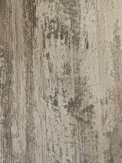 This is a close-up, overhead shot of a textured surface. The surface appears to be made of wood or a wood-like material, with a distressed or weathered finish. The color palette is muted, consisting of shades of light gray, beige, and dark brown. Horizontal lines, characteristic of wood grain, are prominent and vary in thickness and intensity, suggesting a rustic or aged appearance. There are darker, irregular patches and streaks scattered across the surface, giving it a mottled or stained look. Some faint, thin scratch marks are also visible, further contributing to the worn aesthetic. The lighting is even and diffused, with no strong shadows, indicating it might be an indoor setting or an overcast day. The image is abstract in nature, focusing solely on the texture and pattern of the material, and does not depict any people, objects, or a specific scene. No text is visible in the image.