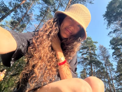 In a forest setting near the city of Barangol, Russia, a woman with long, curly brown hair and a wide-brimmed straw hat is captured in a close-up, low-angle selfie. She is wearing a black t-shirt and a red bracelet on her left wrist, which features some text. A watch with a gold band is also visible on the same wrist. The background consists of tall pine trees against a clear blue sky. In the lower-left corner of the frame, a second individual wearing a yellow jacket and sunglasses can be seen walking away from the camera. The scene appears to be an outdoor recreational or travel setting.