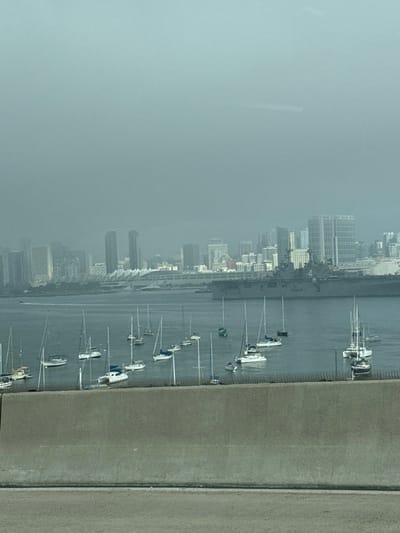 A waterfront cityscape is viewed from an elevated position, likely a bridge or high building. Numerous sailboats rest in the water along the shoreline. The city skyline is partially obscured by a thick haze or fog, muting the colors and details of the buildings. The scene suggests a somewhat overcast or humid day.