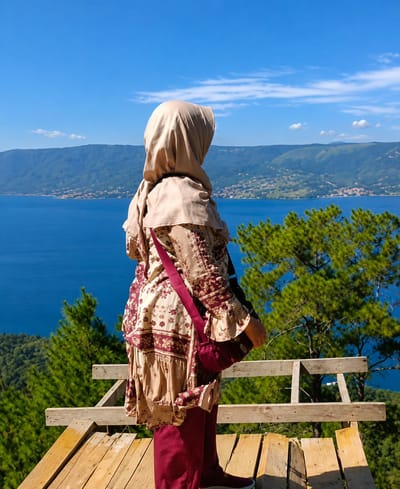 A person wearing a beige hijab and a patterned outfit stands on a wooden platform, looking out at a wide blue lake surrounded by green, rolling hills under a clear blue sky with scattered white clouds. The person is seen from the back, with a maroon bag slung across their body. The platform appears to be a viewpoint, with wooden railings. In the distance, a town is visible nestled along the shore of the lake. This scene is located in Parik Sabungan, Indonesia.