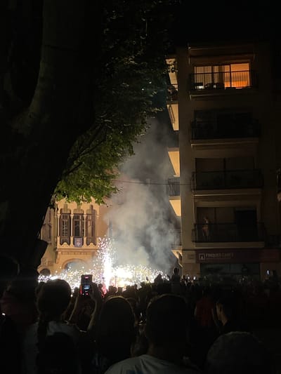 The image captures a nighttime scene in Canet de Mar, Spain, during a fireworks display. A large crowd of people is gathered, many holding up cell phones to record the event. The fireworks are creating a bright, sparkling effect against the dark sky and illuminating the surrounding buildings. In the foreground, the silhouettes of people are visible, with a large tree on the left side of the frame partially obscuring the view.

The architecture of the buildings suggests a Mediterranean style, with balconies and ornate windows. One building on the right has a sign that reads "FINQUES CANET," indicating a real estate agency. Smoke from the fireworks drifts through the air, adding to the atmospheric effect. The lighting is dramatic, with the warm glow from the building windows contrasting with the cool, bright light of the fireworks and the deep shadows of the night. The overall impression is one of excitement and celebration.