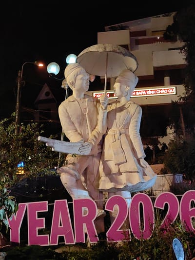 A nighttime outdoor photograph displays a light-colored statue depicting two figures, male and female, located in Sa Pa, Vietnam. The male figure, positioned on the left, wears a long-sleeved shirt and short pants, holding a hollow, cylindrical object resembling a horn or trumpet in his left arm. The female figure, on the right, wears a long-sleeved dress with a tied belt and holds an open, light-colored umbrella above both figures with her left hand. The figures stand close, appearing to face each other with a slight upward gaze, their right hands in proximity, suggesting interaction.
In the immediate foreground, large, freestanding pink letters with white edges spell "YEAR 202"; the final digit is obscured or out of frame to the right. These letters are embedded within dark green foliage. The background features a multi-story building with balconies and a flat roofline, below which a red neon sign displays the Vietnamese text "SAPA KINH CHAO QU". Streetlights with amber and spherical white bulbs provide illumination for the scene, casting light and shadows. A utility pole with wires is visible on the far left. Dark vegetation and other indistinct structures complete the background environment.