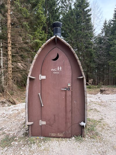 The image shows a public toilet facility located in Kamnik, Slovenia. The toilet is designed to resemble the bow of a ship, constructed from brown wooden planks. The entrance features a semicircular cutout above the door and markings indicating it as a WC (water closet). On the right side of the door, the word "Kazuba" is stenciled in a vertically oriented design. The toilet is situated near a gravel path or parking area, with a backdrop of evergreen trees. A ventilation pipe is visible on top of the facility. The surroundings appear to be a natural setting, likely a park or wooded area near the city. There are no people present in the image.