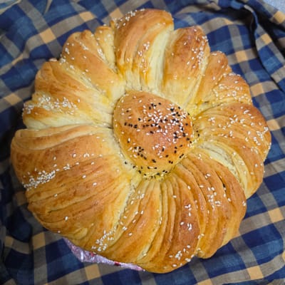 A close-up overhead shot shows a golden-brown, flower-shaped bread sitting on a blue and orange checkered cloth. The bread has a central, rounded portion sprinkled with white and black sesame seeds, surrounded by eight petal-like sections also dusted with white sesame seeds. The petals are formed by layers of dough that have been pulled and twisted. The bread appears to be freshly baked and has a glossy sheen. The background is entirely filled with the checkered cloth, which is slightly wrinkled. The location is not specified in the image.