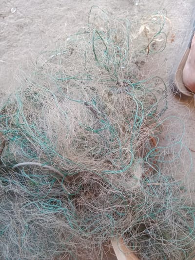 The image displays a close-up, overhead view of a tangled fishing net lying on a sandy surface. The net is composed of thin, interwoven strands, predominantly white and light beige, with some interspersed strands of a light teal or turquoise color. The netting is densely packed and appears somewhat worn, with some strands appearing frayed or knotted. In the upper right portion of the image, a small section of a bare foot, likely a heel and arch, is visible, suggesting a person is present. The sandy ground shows some minor debris, such as small pebbles or fragments. The overall impression is one of discarded or stored fishing equipment in a coastal or sandy environment. The image does not provide any specific visual cues that would allow for the identification of the city of Qobda, Kazakhstan.