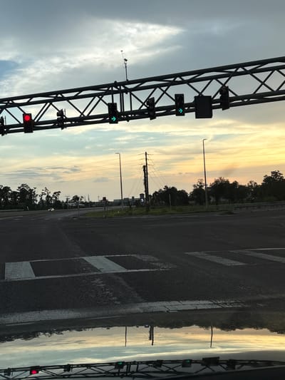The image depicts a street scene, likely a highway or toll plaza, at dusk. The sky displays a gradient of colors, from grey clouds on the left to a soft yellow and orange hue on the right, indicating the sun is setting. The road, wet from recent rain, reflects the sky's colors and the overhead structures.
Dominating the left side of the frame is a large metal gantry, part of an overhead traffic control system. It supports several traffic lights, with a red light illuminated on the lower left and a green arrow pointing upwards on a light further up. Small black boxes are attached to the gantry, possibly housing sensors or cameras. A small antenna-like device is also mounted on the gantry.
The right side of the image shows a paved road with white lane markings. Beyond the road, a line of trees forms a dark silhouette against the colorful sky. Power lines and utility poles are visible in the middle ground. The overall impression is one of a quiet, transitional moment at the end of the day, with the artificial lights of the traffic system contrasting with the natural light of the sunset. No people are visible in the image.