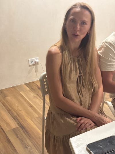 A woman with long blonde hair is sitting at a white table. She is wearing a sleeveless beige dress. Her hands are clasped in her lap. To her right, another person is visible, wearing a white t-shirt. The room has a light-colored wall with an electrical outlet and a power cord plugged in. The floor is made of light brown wooden planks. The scene is indoors, likely in a residential setting, in Verkhniy Yurt, Russia.