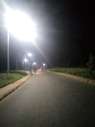 A dimly lit road stretches into the distance under the glow of spaced streetlights. The leftmost light is intensely bright. Two figures walk on the road, their forms indistinct. A vehicle's headlights shine in the background. Grass and low bushes line the left side of the road. A rounded hedge borders the right side. The sky is dark and featureless.