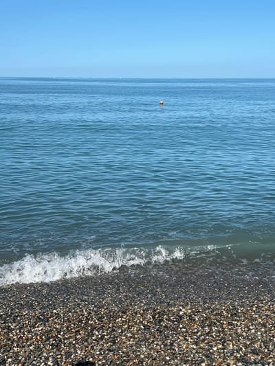 The image depicts a serene coastal scene under a clear blue sky. The foreground is a pebble beach composed of small, multi-colored stones. Gentle waves with white foam are breaking on the shore. The vast expanse of the ocean occupies the majority of the frame, with subtle ripples on its surface. A small, orange buoy floats in the distance, and a hazy horizon line separates the sea from the sky. The image captures a moment of tranquility and natural beauty, likely a view of the Black Sea coast near the city of Prohress, Russia.