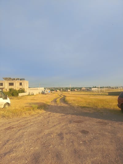 The image depicts an outdoor scene in Yeghvard, Armenia, on a clear day with a pale blue sky, suggesting it is either early morning or late afternoon due to the warm, golden light illuminating the landscape. The foreground is dominated by a dirt road, worn by tire tracks and scattered with pebbles. To the left of the road, a portion of a white van is visible, its wheel and lower body slightly obscured by the camera's angle.
The middle ground features a field of dry, golden grass, typical of a steppe-like environment. A dirt path winds its way through this field, leading towards a collection of buildings in the distance. The most prominent structure is a multi-story building under construction, with visible solar panels on its roof. Adjacent to it are piles of building materials, including bricks and cinder blocks, and what appears to be a partially constructed wall. Further back, more houses and structures form a small settlement, with some featuring pitched roofs. Utility poles with wires crisscross the landscape, indicating the presence of electricity.
On the right side of the image, the rear of a dark-colored car is visible, parked near the edge of the road. The overall impression is one of a developing rural or semi-urban area, with ongoing construction and a sense of open, somewhat arid terrain. The lack of people suggests a quiet moment, perhaps the pause of work or the end of a busy day.