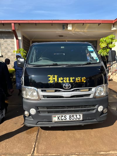 The image shows a black van with the word "Hearse" in gold letters on the front. The van is parked in front of a building. The van has a Kenyan license plate that reads "KCS 853J". The image is taken in front of a house in Githurai, Kenya, with coordinates latitude: -1.179, longitude: 36.914. The scene shows a person in a blue dress and a white head wrap standing by the van, while a person in a black suit stands in front of the van. The van is parked on concrete pavement in front of a building with a stone wall. The building has a red roof and is located on a residential street. The sky is blue and clear. There is a tree in the background behind the van.