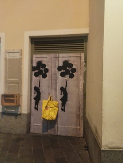 The image depicts a closed double door with stencil art of two silhouettes of girls holding balloons. The left silhouette is on the left door, and the right silhouette is on the right door. A yellow shopping bag hangs from the handle of the left door, positioned below the silhouette of the girl and to the right of her. The doors are set within a building's facade. To the left of the double doors, there is a narrow vertical structure with a light-colored surface and a framed opening. Below this structure, a small wooden shelf unit with two open compartments is mounted on a grey, speckled base. The overall environment appears to be an outdoor urban setting, possibly a street or alleyway, indicated by the pavement in the foreground. The lighting suggests it is either evening or night. This scene is located in Lugano, Switzerland, specifically on Via Francesco Sozzi 6, which is a location known for its street art.