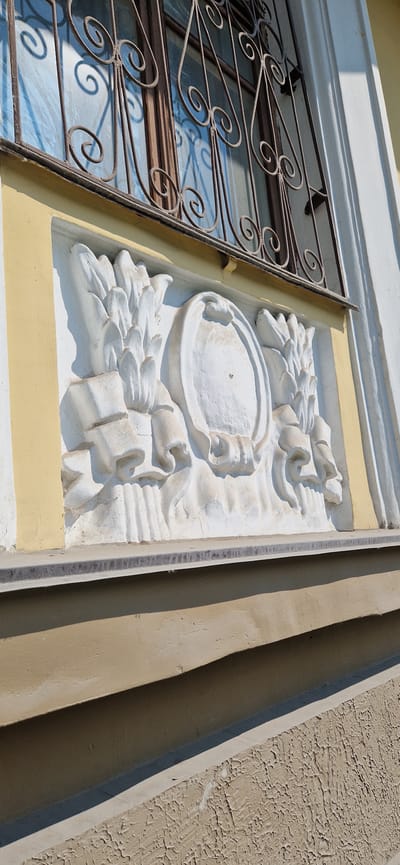 The image shows a close-up, low-angle view of a decorative stucco relief on the exterior wall of a building. The relief is white and appears to be aged, with some visible cracks and imperfections in the stucco. It features a central oval shape framed by what looks like stylized floral or leaf elements on either side. The relief is mounted on a section of the wall painted a pale yellow, which contrasts with the white of the stucco. Above the relief, a portion of a window with ornate wrought-iron bars is visible, casting shadows on the glass behind it. The window frame appears to be made of dark wood. The building's exterior also shows areas painted in lighter off-white and a darker beige, with textured surfaces. The lighting suggests it is daytime, with harsh shadows indicating direct sunlight. The provided context states this image is located in the city of Miass, Russia, but the specific building or context is not visually identifiable from the image itself.