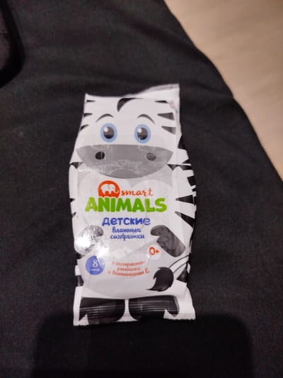 A close-up shot shows a package of wet wipes designed with a cartoon zebra face. The package is white with black stripes and the zebra's face features blue eyes, a black nose, and black eyebrows. The brand name "smart ANIMALS" is prominently displayed in orange and green text. Below this, in blue text, is "детские влажные салфетки," which translates to "children's wet wipes." There is also a circular icon with the number "8" and a plus sign, indicating the age appropriateness of "0+". The wipes are described as containing chamomile extract and vitamin E. The product is placed on a dark, textured surface. No specific location in Mikhailovka, Russia is discernible from the image.