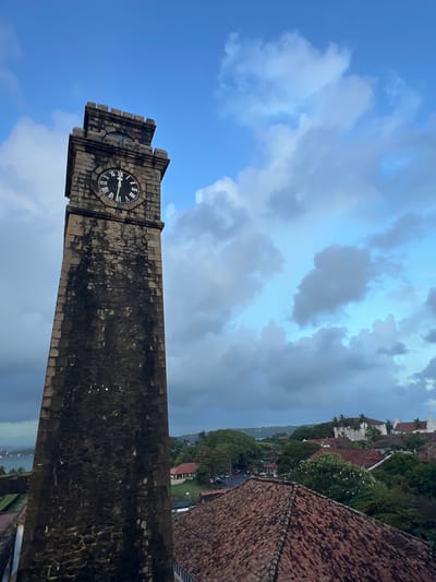 The image depicts a clock tower in Galle, Sri Lanka, against a backdrop of a dynamic sky. The clock tower, constructed of stone, features Roman numerals on its face, indicating a time between 9 and 10 o'clock. The sky is filled with a mix of bright blue patches and dramatic, dark clouds, suggesting variable weather, possibly a storm approaching or passing.
In the background, a cluster of buildings with red-tiled roofs is visible, nestled amongst lush green trees and foliage. The architecture suggests a historical or colonial influence, consistent with Galle's Dutch colonial past. Further in the distance, a body of water can be seen, with hills or a coastline on the horizon.
The overall impression is one of atmospheric beauty, capturing a moment where man-made structures and natural elements converge. The composition, with the clock tower dominating the foreground and the town and sky stretching into the background, creates a sense of depth and scale. The image appears to have been taken during the late afternoon or early evening, given the quality of the light and the cloud formations. There is no visible text in the image.