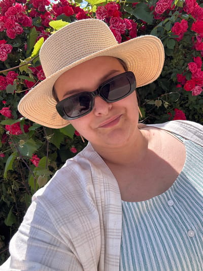 A woman wearing a straw hat, sunglasses, a white cardigan, and a striped top poses for a selfie in front of a blooming rose bush. The image appears to have been taken outdoors, possibly in a garden or park, under bright sunlight. The background is filled with vibrant red roses and green foliage, suggesting a warm, sunny day. The woman is looking towards the camera with a slight smile. The location mentioned, Добровское сельское поселение, Ukraine, is a rural administrative division. However, there are no specific visual cues in the image that definitively confirm this location.
