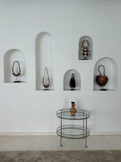 The image shows a display of artifacts on a white wall, possibly in Ghadeer Al Tayr, United Arab Emirates. Six recessed niches contain various objects resembling tribal or ethnographic jewelry and adornments. The items include beaded necklaces, feathered objects, and possibly ceremonial ornaments. To the right of the niches is a metal and glass circular shelving unit holding a decorative vase or urn. The overall aesthetic is modern and minimalist, highlighting the artifacts as cultural or artistic objects. The wall is part of an interior space, with a section of tiled or stone wall visible to the right of the shelving unit.