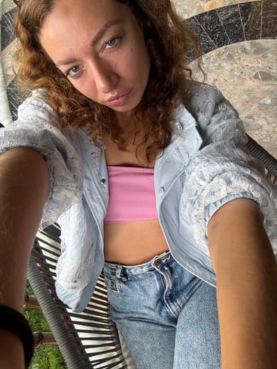 A young adult female is depicted taking a selfie, looking directly into the camera with her head slightly tilted to the left and lips subtly pursed, exhibiting a serious expression. She has long, wavy to curly, reddish-brown hair, parted on the left side. Her eyes are light-colored, appearing greenish-blue. Her skin is fair, with visible minor blemishes or freckles on her face.
She is wearing a pink strapless bandeau-style crop top, which is partially covered by a light blue bomber-style jacket. The jacket features a zippered front closure and snap buttons on the collar and cuffs. Its sleeves and portions of the back are made of a translucent white lace material. She is also wearing light blue denim jeans with a visible button and a partially unzipped fly. One arm, appearing tan and lightly haired, extends into the foreground from the lower left, holding the camera device out of frame.
She is seated in an outdoor chair with a dark gray woven or corded backrest and a partial armrest visible on the left. The background features a distinctive tiled floor with a geometric pattern composed of various shades of gray, black, and beige stone or marble, including curved and radial elements. Beyond the chair and to the left, green foliage from plants or trees is visible, indicating an outdoor setting. The scene is well-lit by natural light. The specific geographic location of Púng Luông, Vietnam, cannot be verified from the image content.