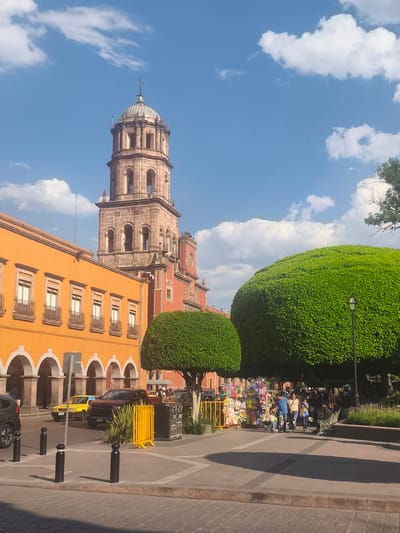 This image captures a sunny day in Querétaro, Mexico, featuring a prominent church tower and colonial architecture. The foreground shows a pedestrian area with large, meticulously trimmed trees, and vendors with their wares. People, including families, are walking and browsing. Behind them, vehicles, including a taxi, are parked along the street. The architecture consists of colorful buildings, primarily in shades of orange and red, with arches and windows. The sky is bright blue with scattered clouds.