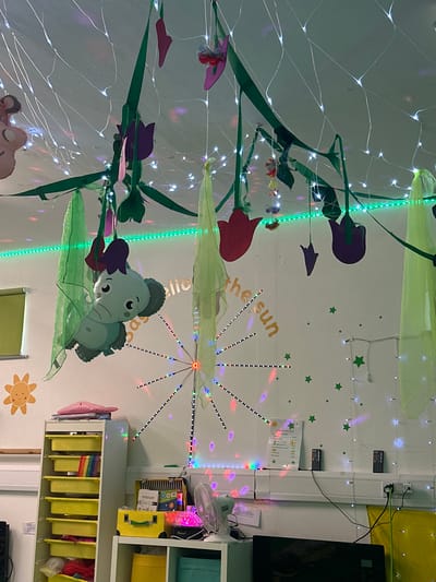 The image depicts a wall decorated with playful, childlike elements, possibly in a classroom or a child's room. The decor includes a string of white fairy lights, green streamers, and several paper cutouts of purple and red flowers, along with a blue elephant character. Green star stickers are scattered across the wall, and a sun graphic is visible in the lower right corner.

A section of the wall features a light fixture with colorful, blinking LED lights arranged in a radiating pattern. Below this, the words "the sun" are written in a gold, textured font. Two remote controls are attached to the wall near the top right, and a small paper with text, possibly a price list, is also affixed to the wall.

In the background, part of a white shelving unit is visible, containing yellow storage bins and some other items. A black monitor or television screen is mounted on the right side of the wall. The overall impression is one of a cheerful and whimsical environment, likely designed to engage young children. There are no people visible in the image. The lighting suggests it may be daytime indoors, or that artificial lights are being used for decoration.