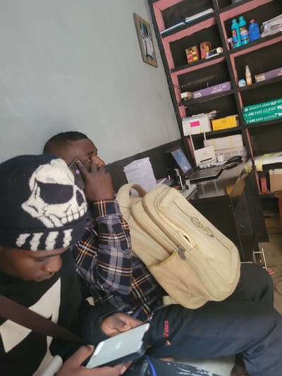 In Bokkos, Nigeria, a scene unfolds within an office or shop. Two individuals are present. The foreground features a person wearing a black beanie with a white skull design and a black sweatshirt. This individual is looking down at a silver tablet, holding it with both hands. To their right, a second person, facing to the right of the frame, is on the phone. This person is wearing a plaid shirt with orange, black, and white checkered patterns, and is holding a dark-colored phone to their right ear. A tan messenger bag rests on their lap, draped over their knees, with a portion of the bag visible.
The environment includes a desk area behind the individuals. On the desk, a laptop is open and powered on, displaying a screen with a blue background and graphical elements. A Wi-Fi router, a black phone, a stapler, and other office supplies are also on the desk. Behind the desk, shelves are filled with various items, including stacks of paper, boxes, bottles of cleaning supplies, and what appear to be reams of copier paper. A framed picture is visible on the wall to the left of the shelves. The lighting suggests an indoor setting, possibly illuminated by overhead lights or natural light from an unseen source. The overall impression is of a functional, perhaps slightly cluttered, workspace.