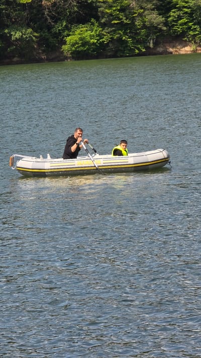 Two people, an adult male and a boy, are in an inflatable boat on a body of water. The adult male is in the rear of the boat, rowing with an oar. He is wearing a black hoodie. The boy is in the front of the boat and is wearing a bright yellow life vest. The boat has "MARINER" written on its side. The background shows a forested shoreline. The scene appears to be on the Pasarel Reservoir, located near the village of Dolni Pasarel, Bulgaria. The water is rippled, suggesting a gentle breeze.