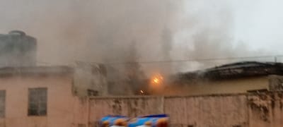 The image captures an outdoor scene dominated by an active fire producing a vast amount of smoke. In the foreground, a light-colored, possibly concrete or masonry, wall or fence spans horizontally across the lower part of the frame, showing some signs of wear and discoloration. At the very bottom of the image, partially obscured by the fence, two blue objects, possibly packages or bags with orange and yellow designs, are visible.
To the left in the mid-ground, a building with light-colored, possibly pinkish-beige, walls is visible. It features at least two dark-paned rectangular windows. On the roof of this building, a dark-colored water tank or similar utility container is present, partially shrouded by smoke.
Towards the center and right of the mid-ground, a bright, intense orange-yellow glow indicates significant flames, serving as the focal point of the fire. This fiery glow is almost entirely engulfed by massive plumes of thick, greyish-white smoke that rise and spread across the entire upper half of the image, obscuring the sky and any background details beyond the immediate structures. The smoke creates a hazy, dim atmosphere throughout the scene. Faint, thin dark lines, possibly overhead wires or cables, are visible stretching horizontally across the mid-ground, partially disappearing into the smoke.
The lighting suggests either an early morning, late evening, or heavily overcast daytime, intensified by the dense smoke. The overall scene conveys an emergency situation involving a fire, with extensive smoke production. No discernible text is visible within the image.