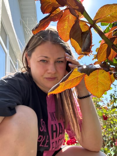 In Uyutnoye, Ukraine, on a sunny day, a woman with long brown hair is partially obscured by a rose bush with reddish-orange leaves. She is wearing a black t-shirt with pink lettering and is crouching down. Her right hand is touching her head, and her left arm is resting on her leg. The background features a white building and a bright blue sky. Red roses and green foliage are visible in the background.