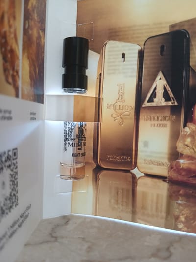 The media file displays a close-up view of a perfume sample or miniature bottle. The bottle is clear glass with a black cap and is positioned horizontally. Visible text on the bottle and packaging includes "PACO RABANNE 1 MILLION", "NIGHT ELIXIR", "MADE IN FRANCE", and "Barcelona London New York Tokyo". The background shows a gold-colored, textured surface, possibly representing gold bars or a similar luxurious material, with a blurry, abstract image of golden elements at the top and bottom. The overall aesthetic suggests a product related to the Paco Rabanne "1 Million" fragrance line, emphasizing luxury and opulence. The scene is likely a product display or personal collection. There are no people or animals visible. The lighting is bright, suggesting an indoor setting during the day.
