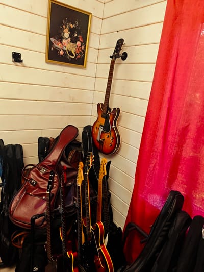 A cluttered corner of a room with light-colored, horizontal wood paneling on the walls. A framed painting of flowers hangs on the wall, above a black metal guitar wall mount (unused). An electric bass guitar hangs from another wall mount. A large, red fabric partially obscures the right side of the frame. On the floor are multiple electric guitars, some in cases, some without. A large, brown leather guitar case sits prominently among the guitar cases. The location is described as being in the city of Свистуха (кузяевский С/о), Russia, but there are no identifiable elements from the image to support that claim. No people or other actions or interactions are visible in the image.
