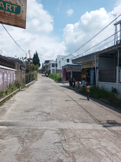 The image depicts a street scene, likely in a residential or mixed-use area. The road is paved but shows signs of wear and tear. On either side of the road, there are buildings. On the right side of the road, several children are standing near a shop that appears to be a laundry. A motorcycle is parked nearby. There are power lines overhead. On the left side of the road, there is a fence topped with barbed wire. There is a sign in the upper left corner with the word "ART" on it. The sky is partly cloudy. The image appears to be taken in the daytime. The location is unknown but not Zeyathiri District, Myanmar. The text on the sign indicates the location is likely Indonesia.